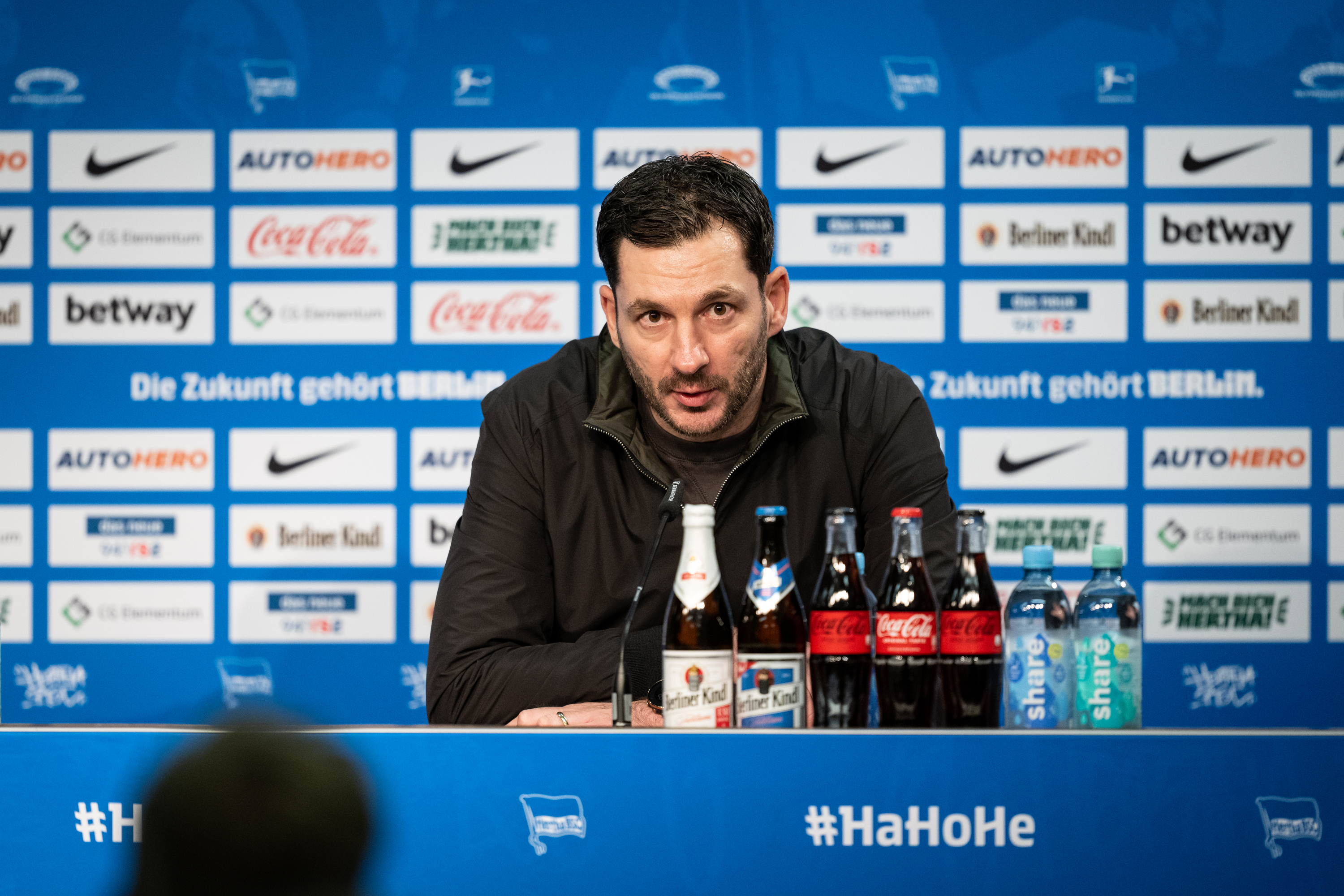 Sandro Schwarz sitzt bei einer Pressekonferenz auf dem Podium und spricht.