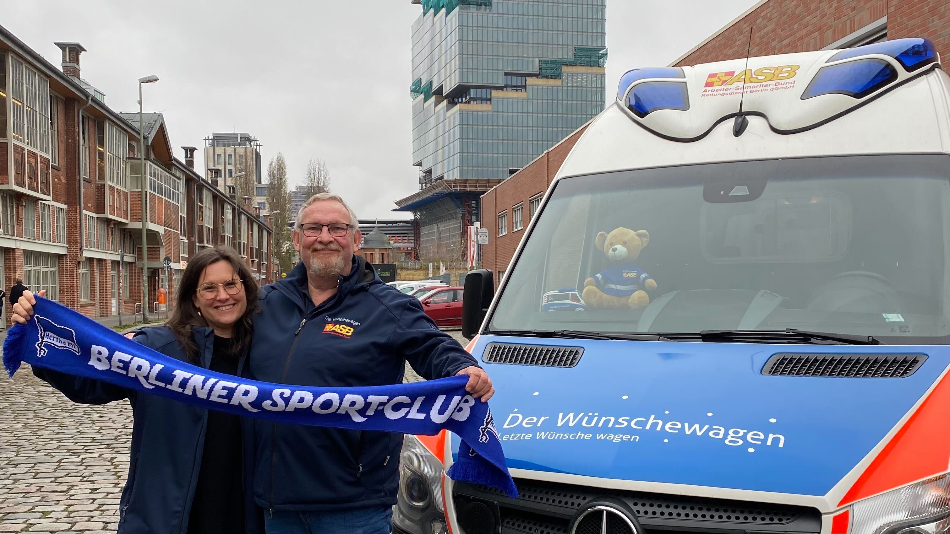 Jörg Bredow und Ann-Brit Keck stehen vor dem Wünschewagen und halten einen Hertha-Schal.