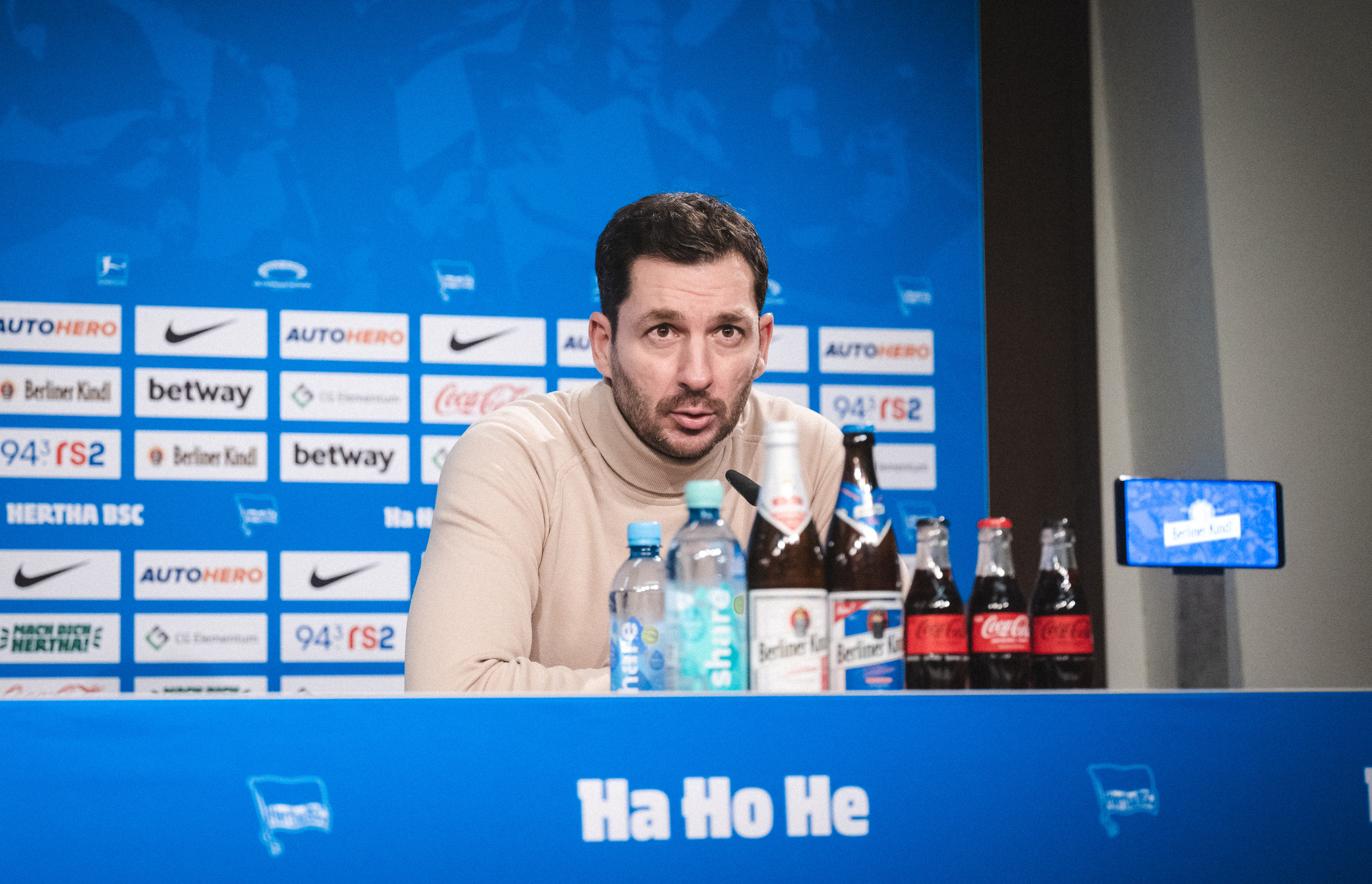 Sandro Schwarz sitzt auf dem Podium und blickt in die Kamera.