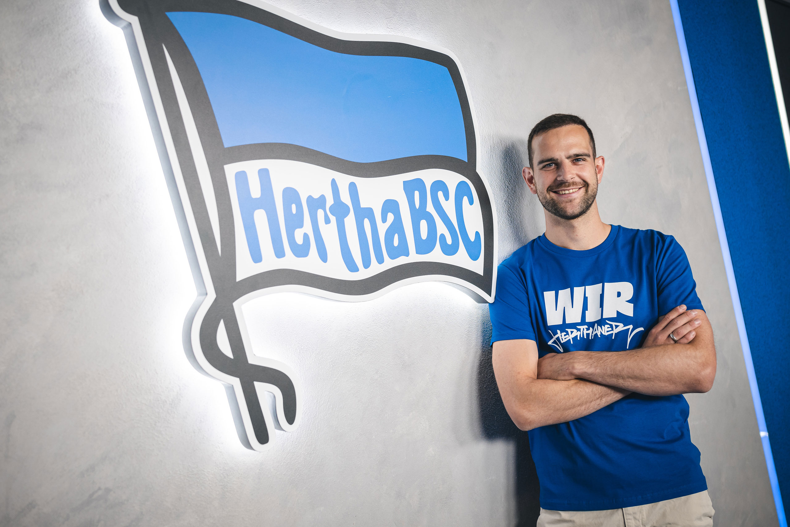 Rückkehrer Marius Gersbeck posiert mit einem Lächeln neben der Hertha-Fahne.