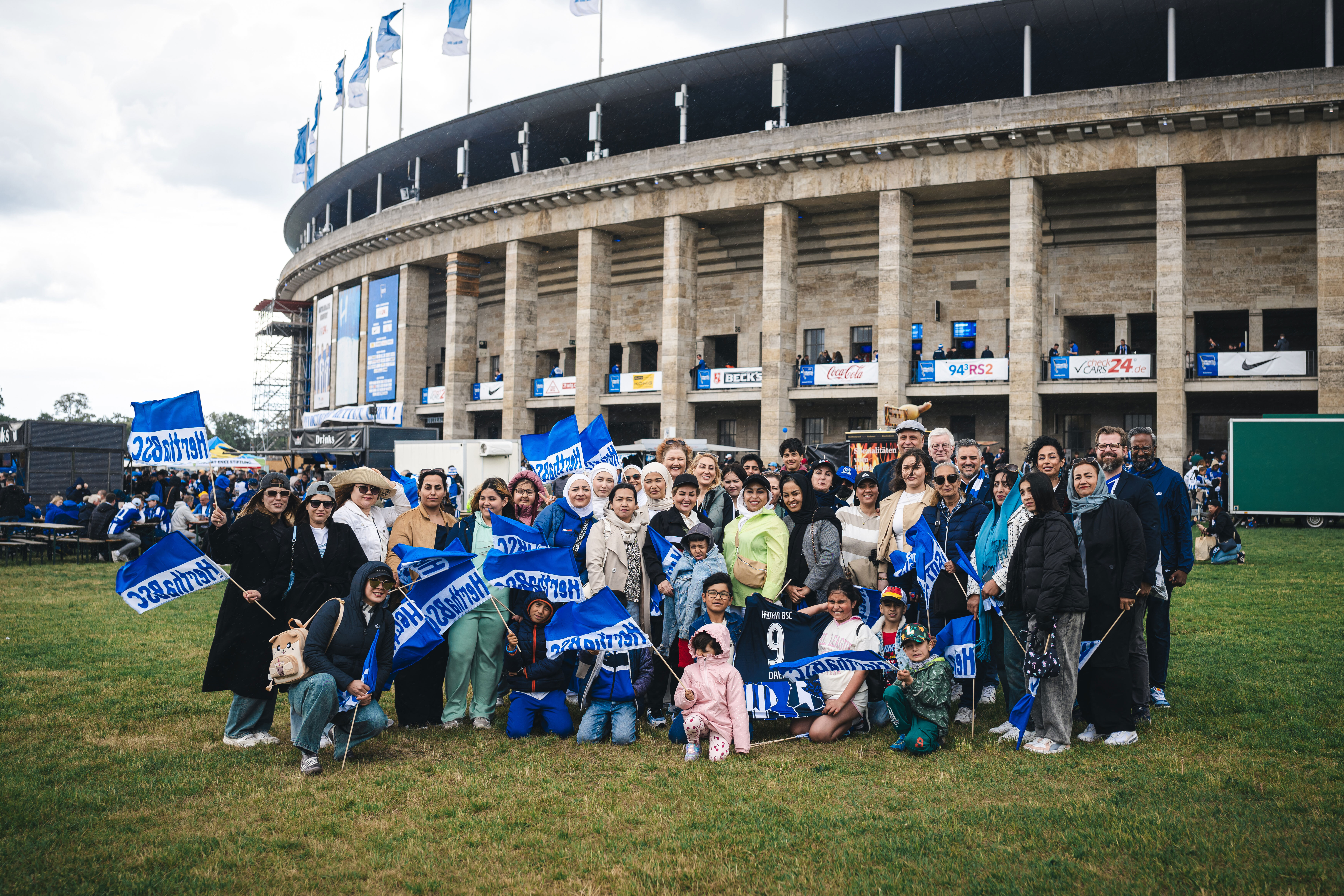 Die Gruppe von Frauen und Kindern mit einigen Präsidiumsmitgliedern vorm Olympiastadion. Sie lachen in die Kamera und halten Hertha BSC Fahnen hoch.