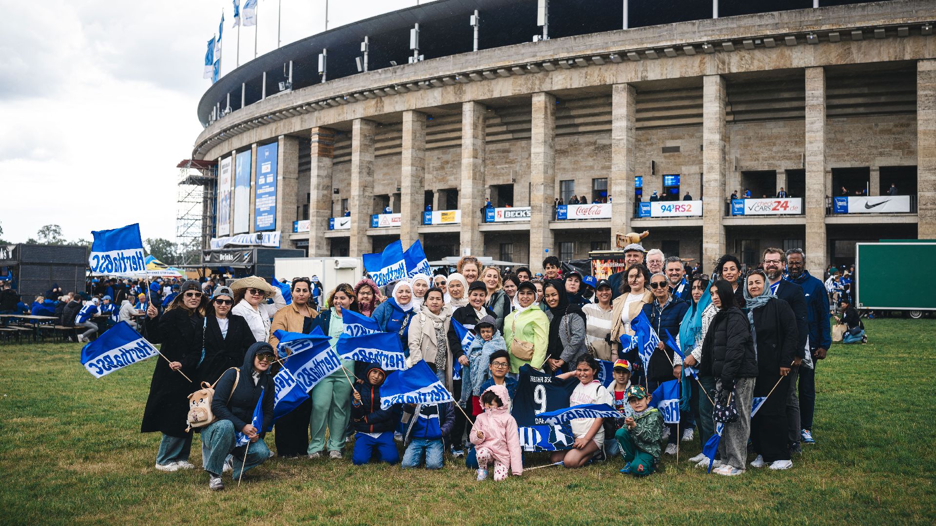 Die Gruppe von Frauen und Kindern mit einigen Präsidiumsmitgliedern vorm Olympiastadion. Sie lachen in die Kamera und halten Hertha BSC Fahnen hoch.