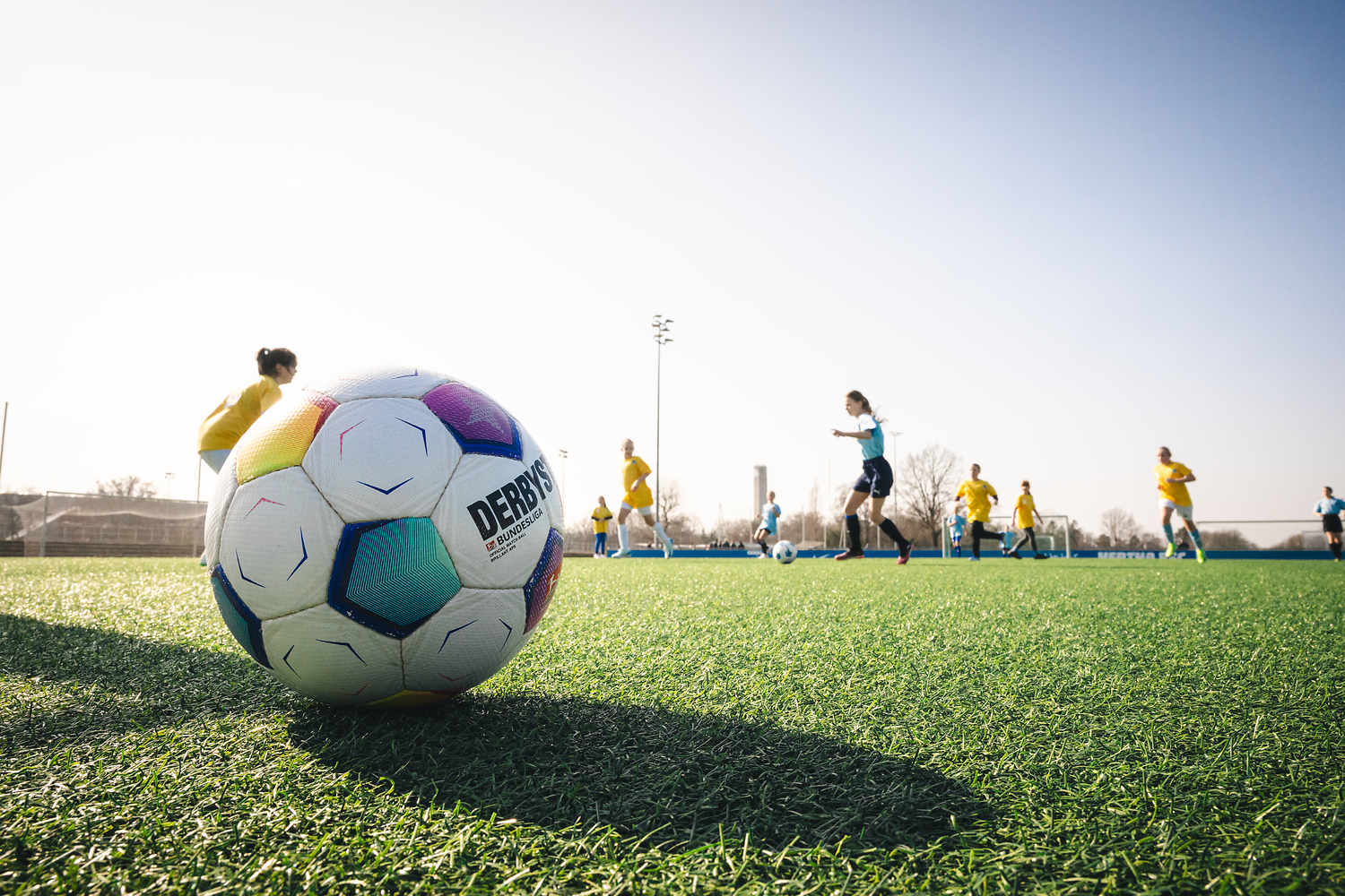 Im Vordergrund liegt ein Ball auf dem Rasen, im Hintergrund spielen Mädchen.