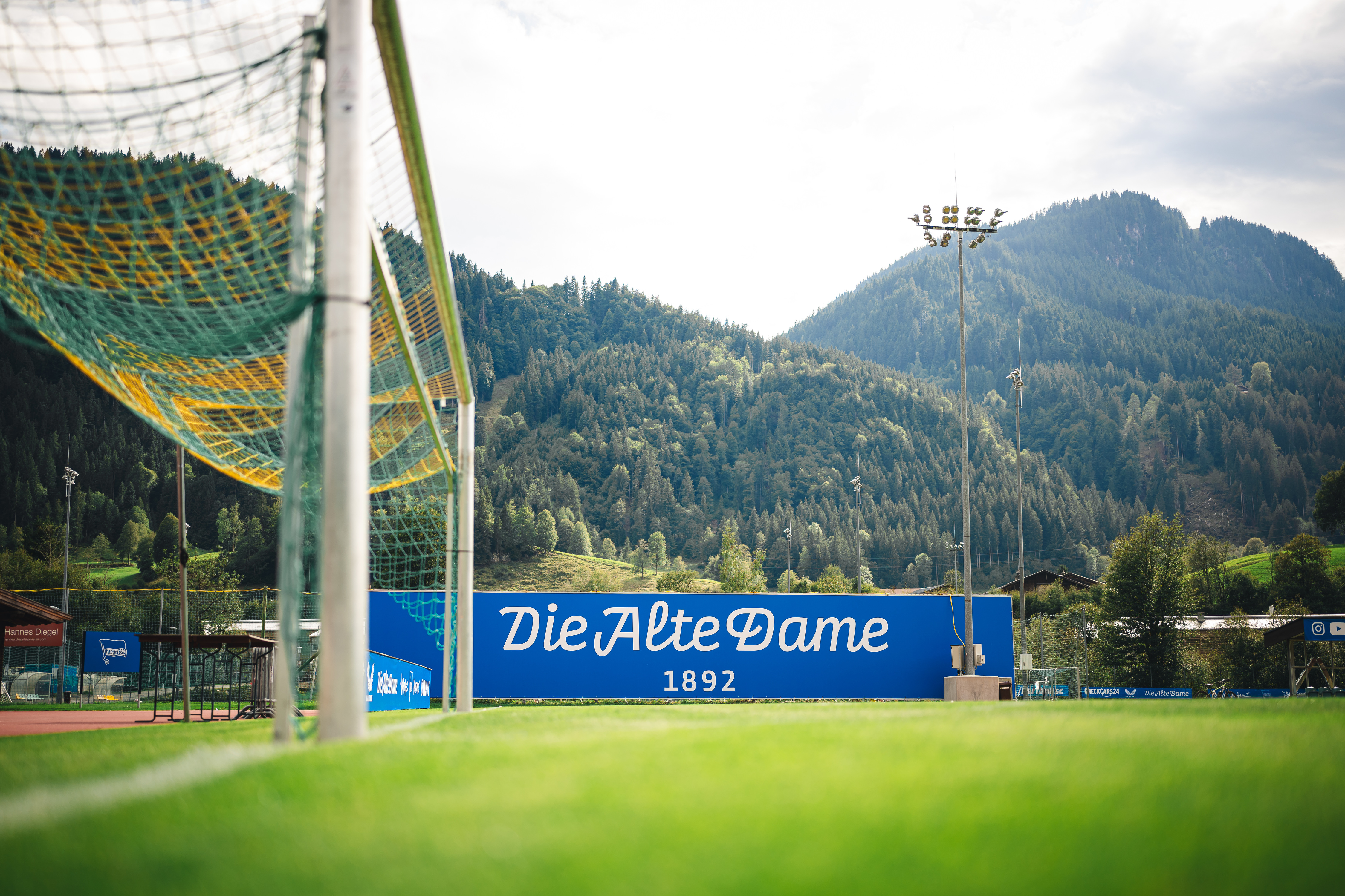 Ein blau-weißer Schriftzug auf dem Trainingsplatz vor einem Berg im Hintergrund.