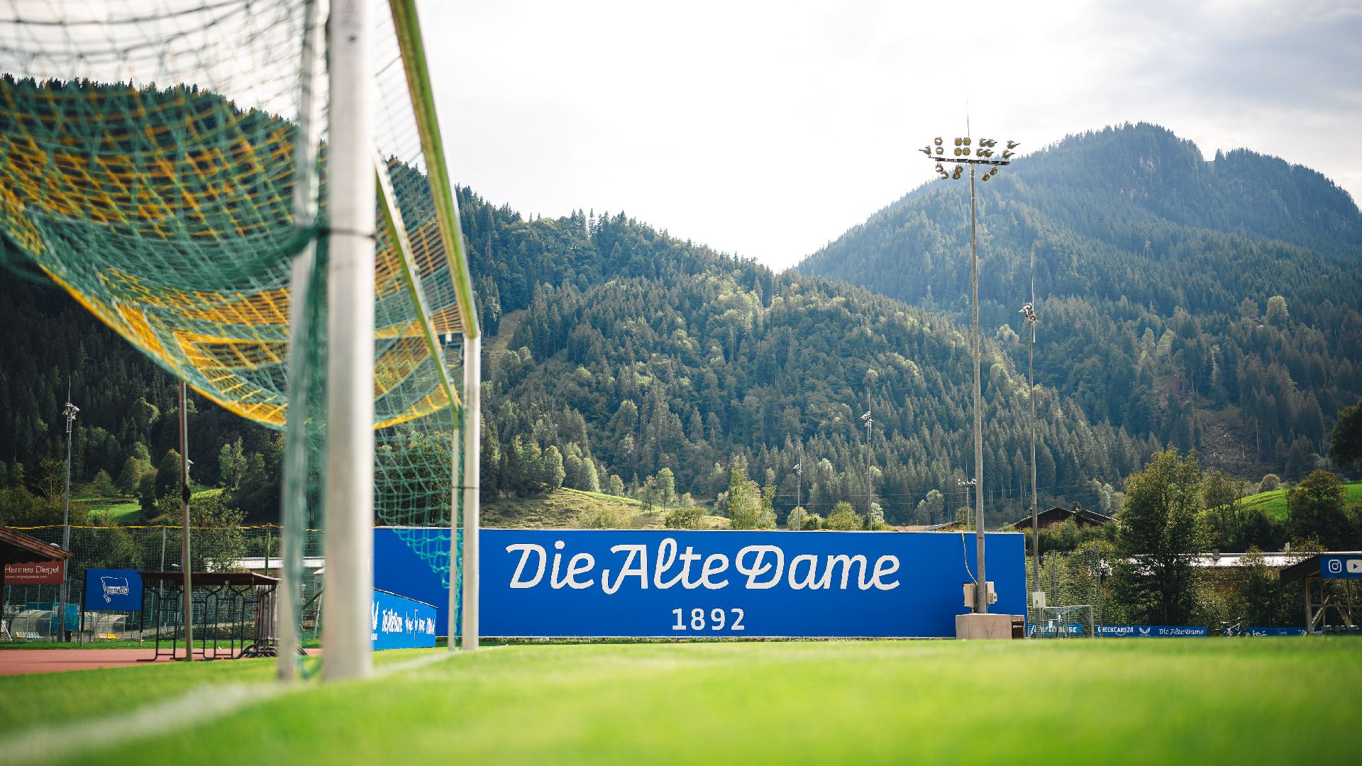 Ein blau-weißer Schriftzug auf dem Trainingsplatz vor einem Berg im Hintergrund.