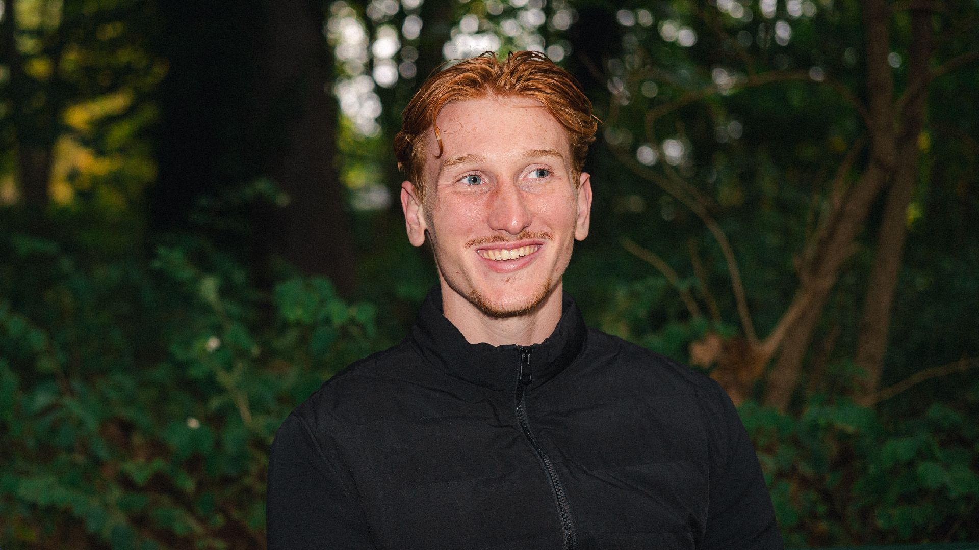 Konstantin Heide sitzt auf einer Bank im Wald und lacht.